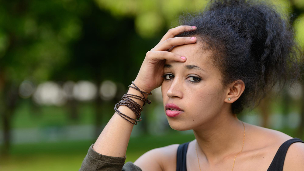woman looking into the camera with hands on head with neutral expression, in a park
